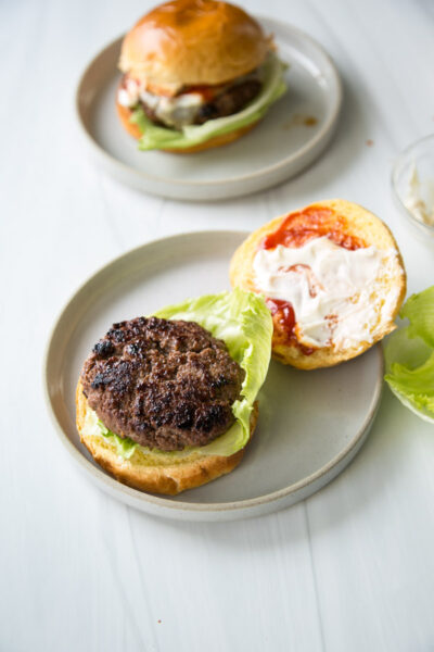 An open-faced, gluten-free hamburger on a plate with a beef patty and lettuce, next to the top bun spread with ketchup and mayonnaise. Another assembled burger is in the background on a separate plate.