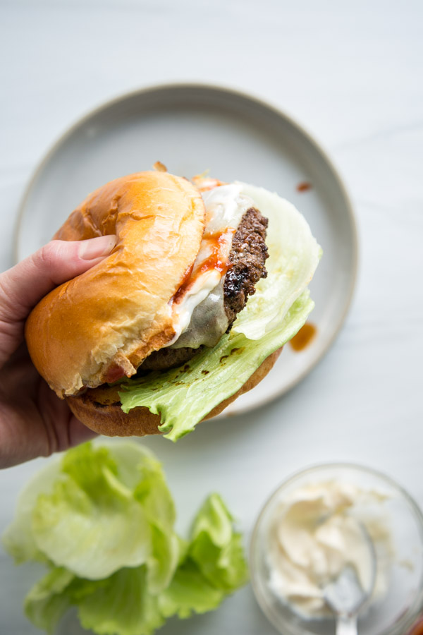 A hand holds a gluten-free hamburger with lettuce, a beef patty, melted cheese, sauce, and a brioche bun above a plate. Nearby are loose lettuce leaves and a bowl of mayonnaise with a spoon.