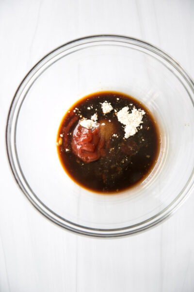 A clear glass bowl containing a mixture of dark soy sauce, ketchup, white powdered seasoning, and other gluten-free sauce ingredients on a white surface.