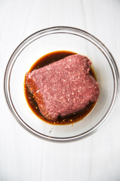 Raw ground beef sits in a clear glass bowl with a dark, gluten-free marinade, perfect for pairing with almond flour sides on a white marble surface.