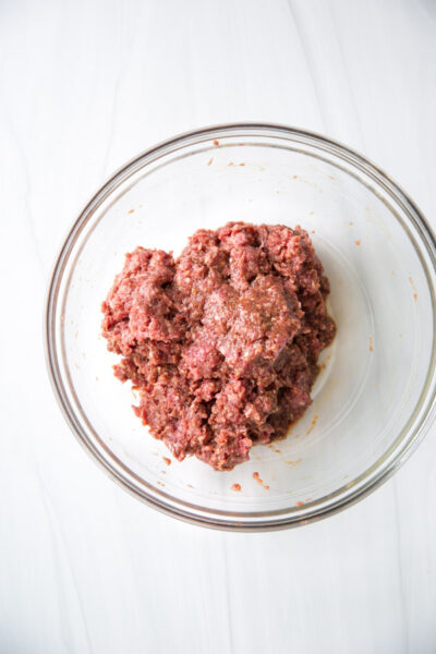 Ground meat mixed with seasonings is shaped like a heart inside a clear glass bowl, placed on a white surface—perfect for creating gluten-free dishes using fresh ingredients.
