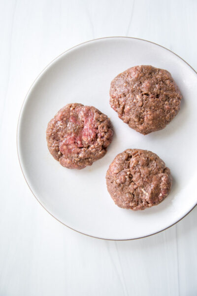 Three raw hamburger patties placed on a white plate, viewed from above on a light-colored surface—perfect for pairing with a gluten-free side or complementing an almond flour-based dish.