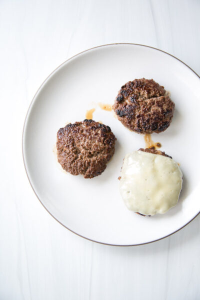 Three cooked, gluten-free beef burger patties on a white plate; two are plain and one is topped with melted cheese. The plate is set on a white surface.