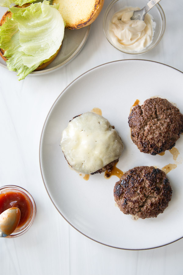 Three cooked burger patties, one topped with melted cheese, are on a white plate. Nearby are a gluten-free bun with lettuce, a bowl of mayo, and a dish of ketchup with a spoon.