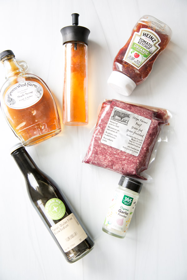 A bottle of apple cider vinegar, a squeeze bottle of oil, gluten-free ketchup, a package of ground beef, Worcestershire sauce, and garlic powder arranged on a white surface.