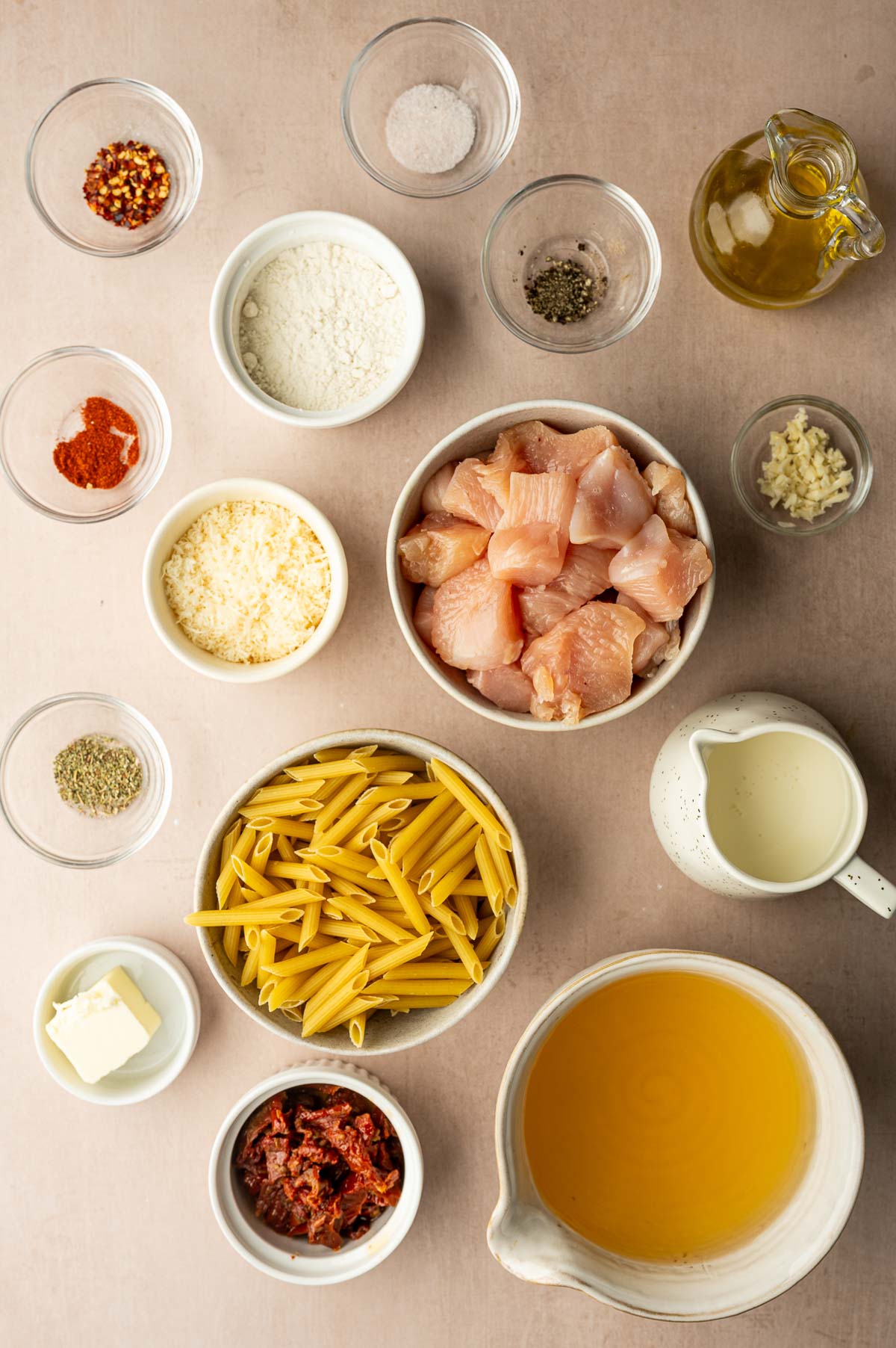 Various ingredients are arranged in bowls on a countertop, including diced chicken, penne pasta, sun-dried tomatoes, broth, seasonings, parmesan cheese, flour, garlic, butter, milk, and oil.
