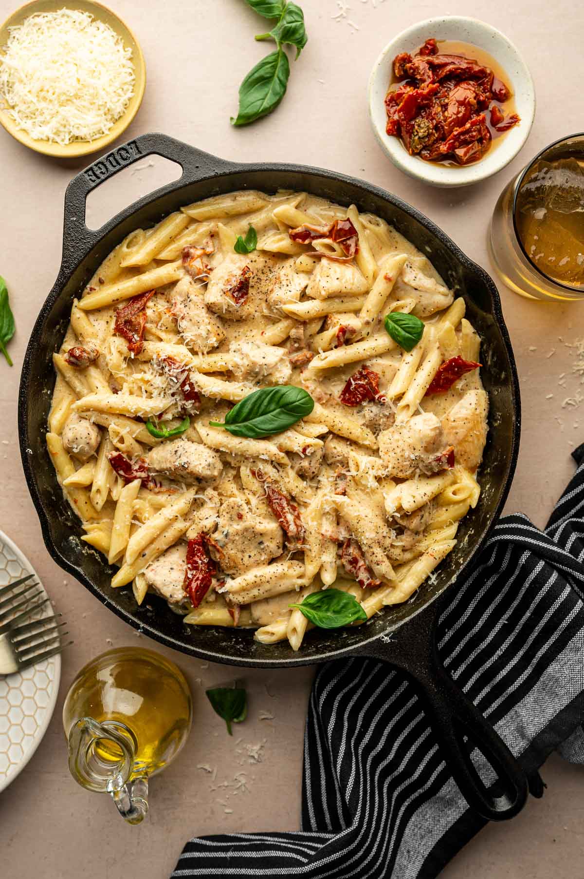 A skillet filled with creamy marry me chicken pasta with penne pasta, sliced chicken, sun-dried tomatoes, and basil, surrounded by plates, grated cheese, olive oil, and a glass of drink.