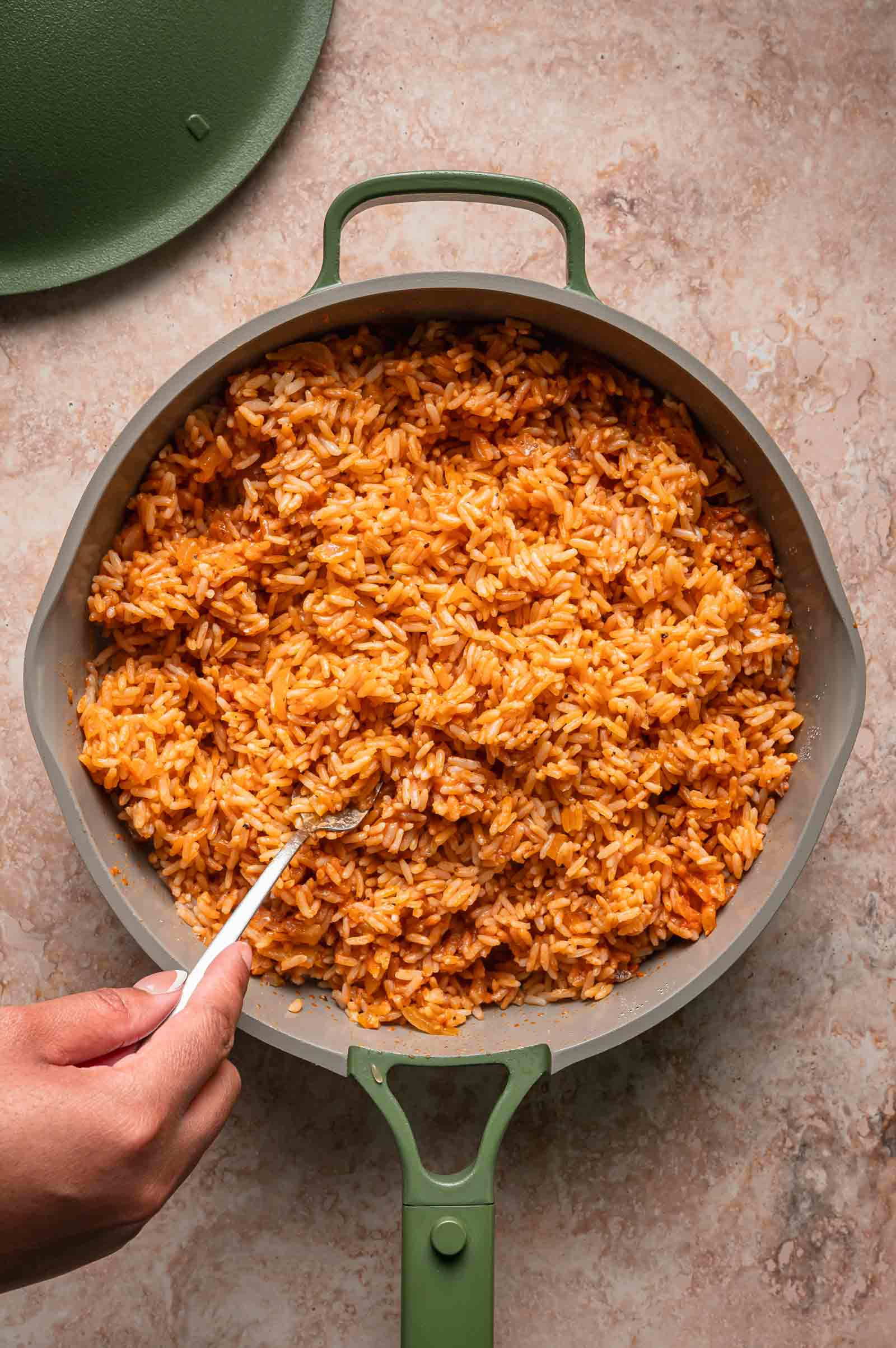 Overhead view of a pan filled with cooked, Spanish rice. A hand is holding a spoon, ready to serve or stir the rice.