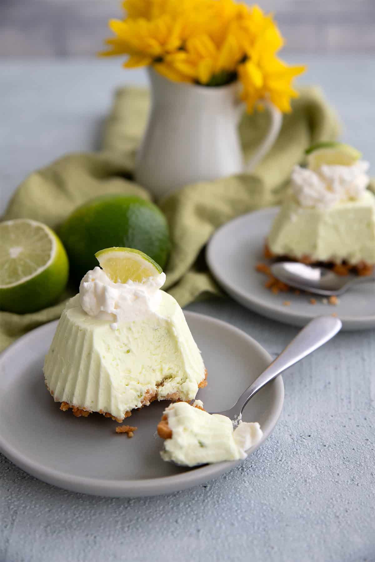 Two mini frozen key lime pies on small gray plates with spoonfuls taken out of them, and a small vase of yellow flowers in the background.