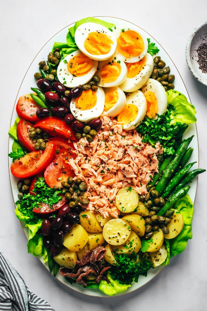 A salmon nicoise salad with salad greens, green beans, new potatoes, tomatoes, hard boiled eggs, olives, capers, salmon and herbs on an oval platter.
