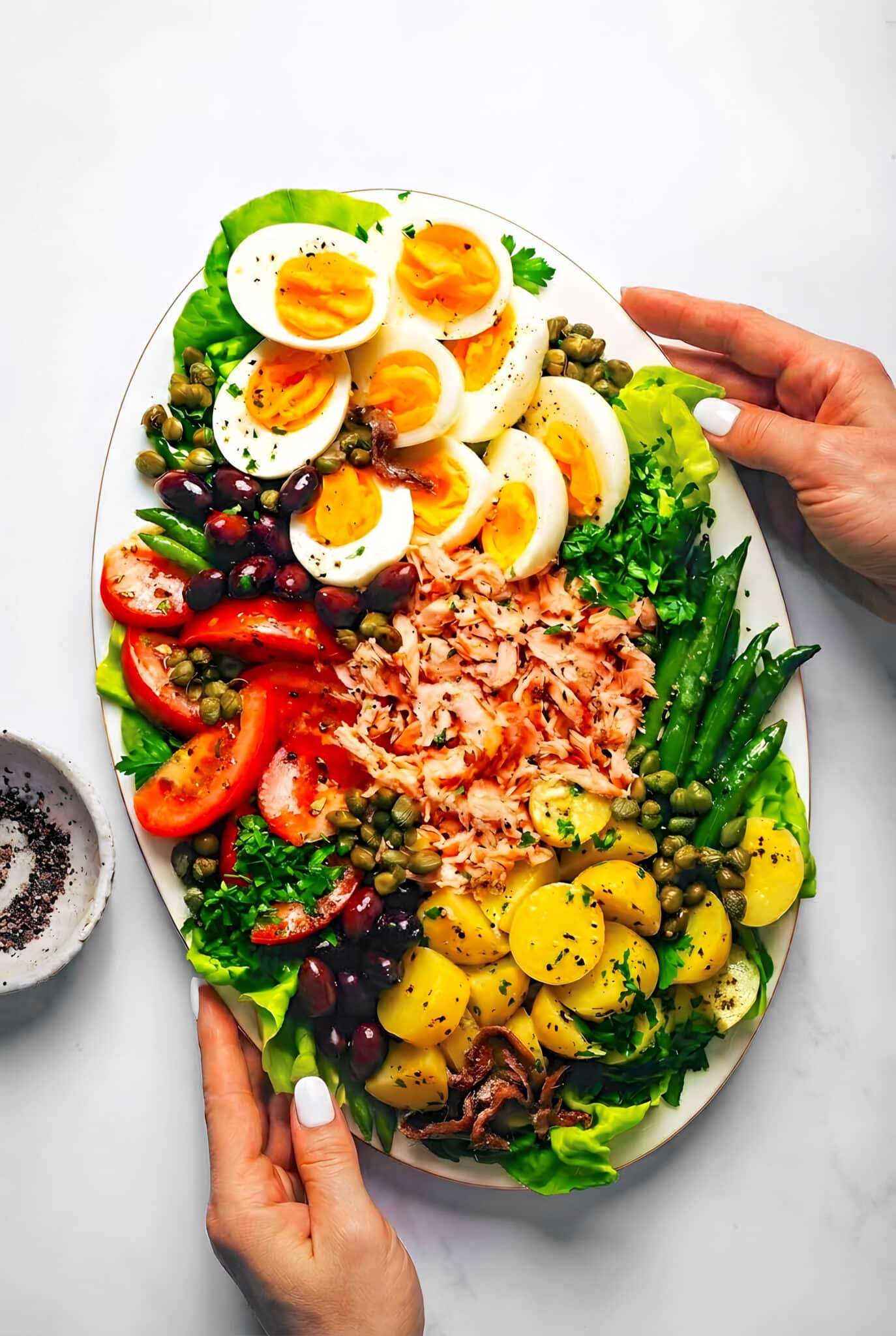 A woman's hands holding a salmon nicoise salad with salad greens, green beans, new potatoes, tomatoes, hard boiled eggs, olives, capers, salmon and herbs on an oval platter.