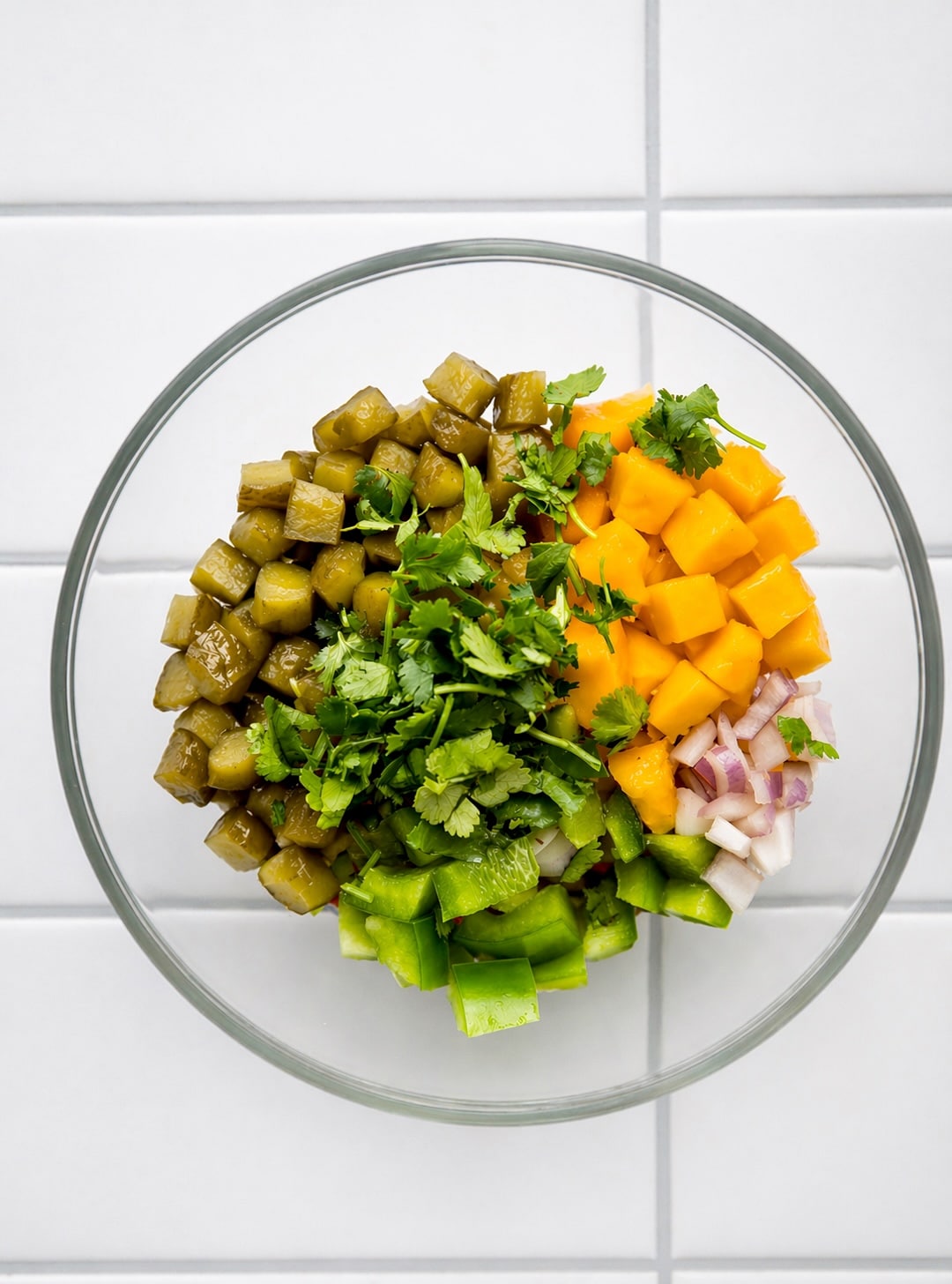 Chopped pickles, mango, green pepper, red onion, and cilantro arranged in a glass bowl before mixing.