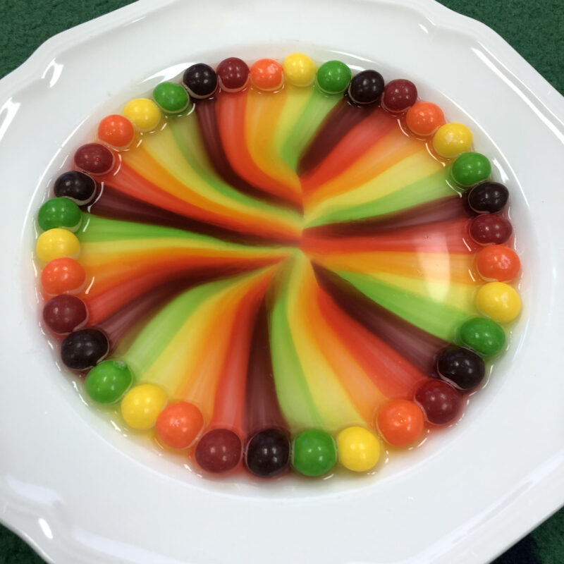 an image of skittles in a shallow dish with water and their colors are combining to make a rainbow