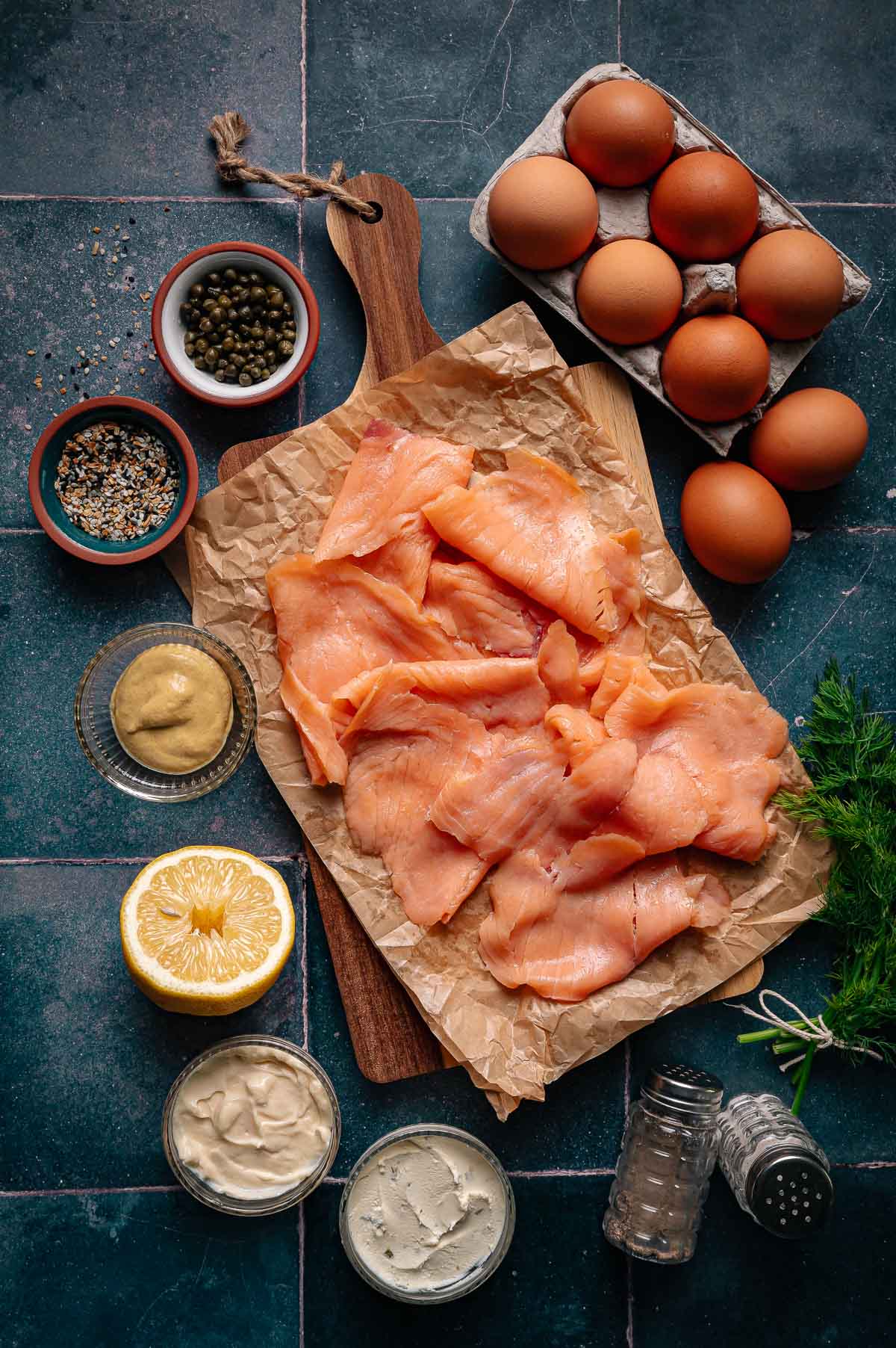 Sliced smoked salmon on parchment paper with eggs, lemon, mustard, mayonnaise, pepper, capers, fresh dill, and seasonings on a dark surface.