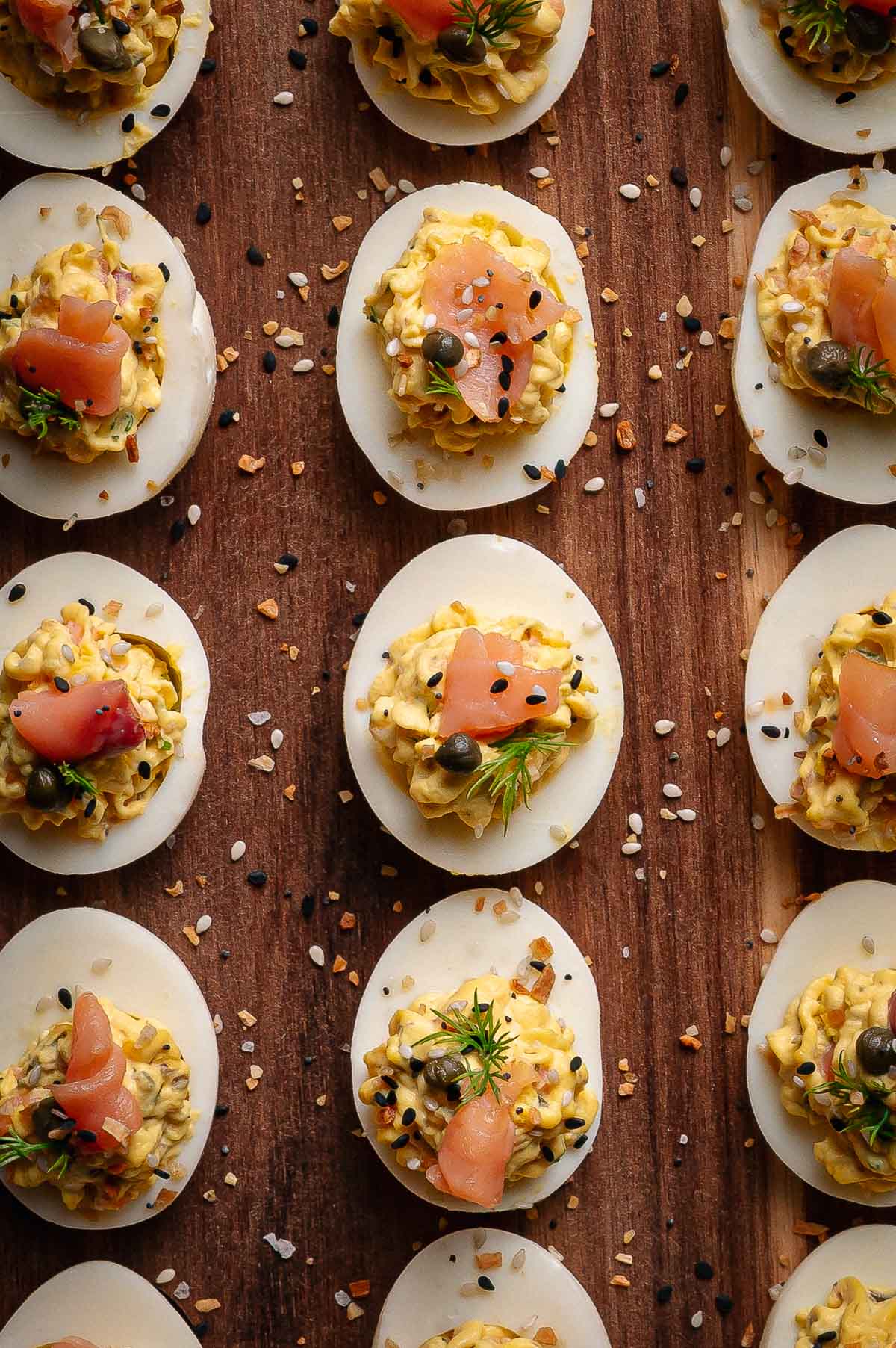 Halved hard-boiled eggs filled with a creamy smoked salmon filling, garnished with herbs, capers, seasoning, and smoked salmon, arranged on a wooden surface.