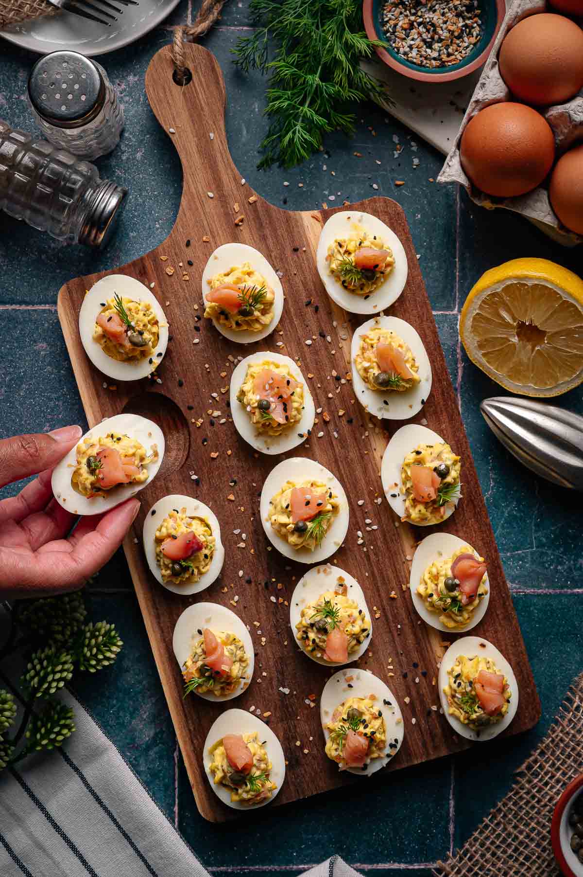 A wooden serving board with several deviled eggs topped with smoked salmon, herbs, and seeds; a hand holds one egg. Eggs, lemon, and seasoning are nearby on a dark countertop.