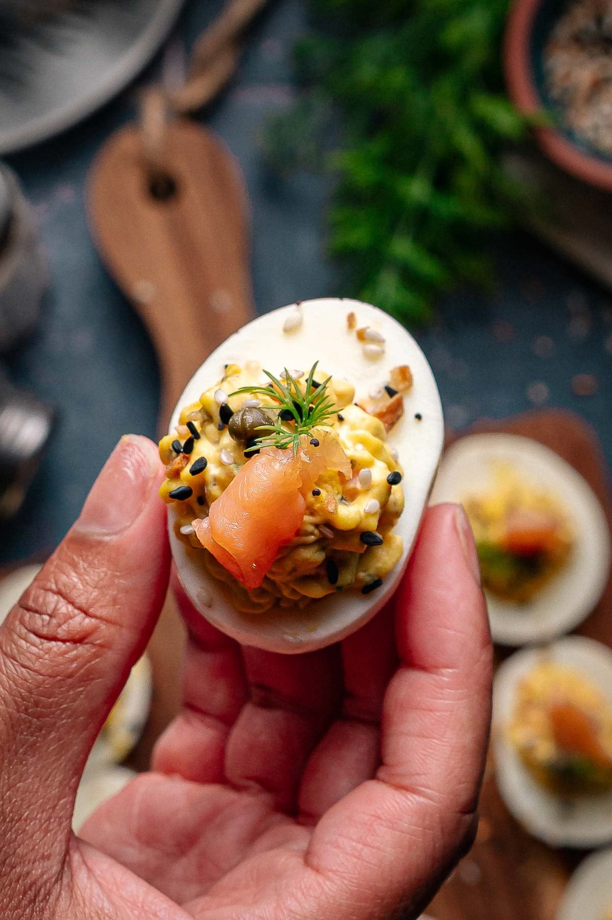 A hand holds a deviled egg topped with smoked salmon, black sesame seeds, dill and capers.