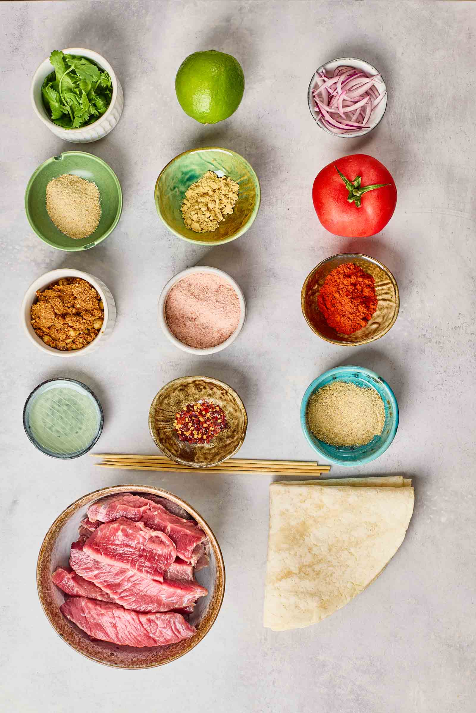 Various ingredients for suya skewers are neatly arranged on a light surface, including raw beef slices, spices, tomato, red onion, lime, greens, a flatbread, and small bowls of powders and seasonings.