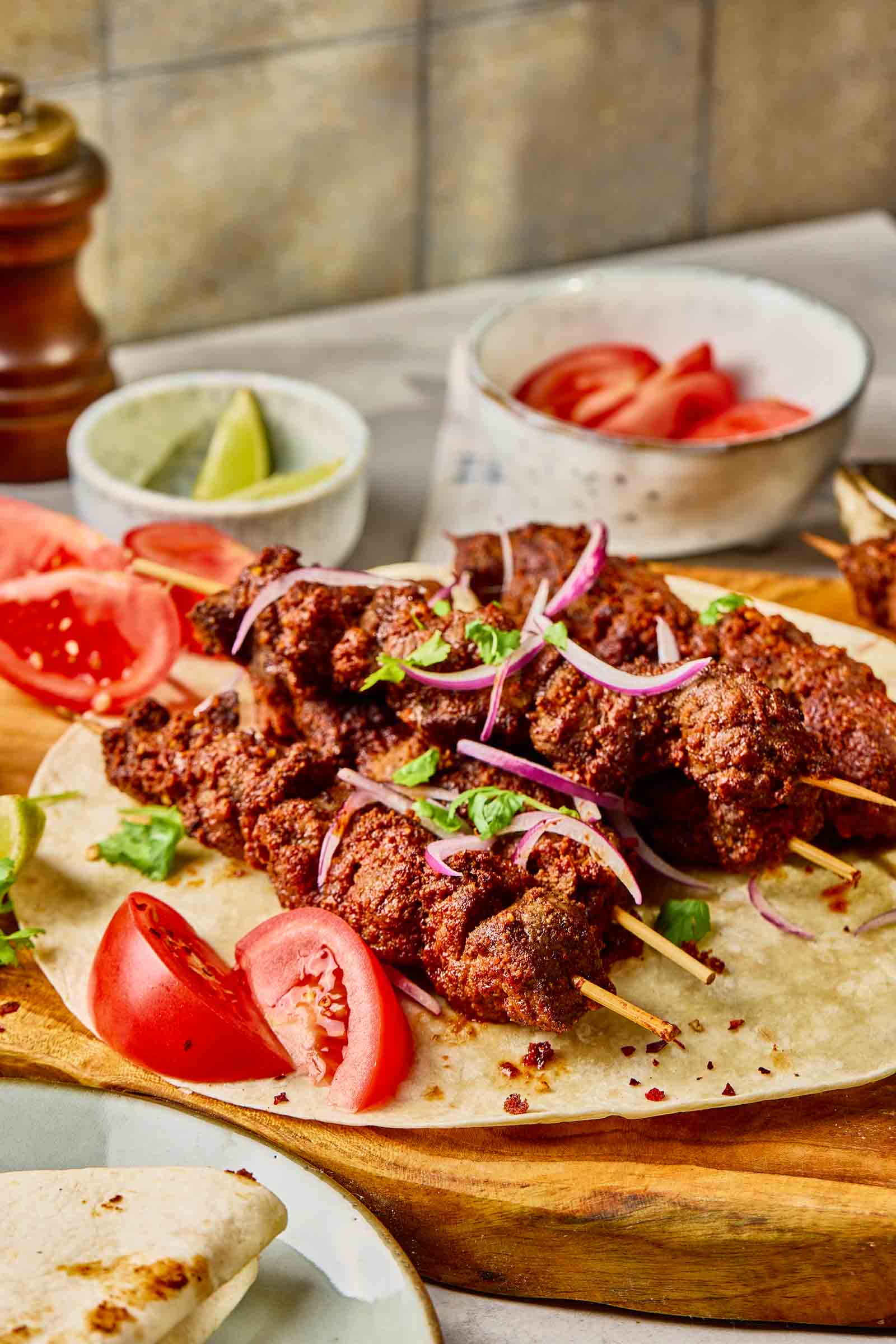 Skewers of seasoned grilled meat served on flatbread, garnished with sliced red onion, tomato wedges, and herbs, with lime wedges and a bowl of sliced tomatoes in the background.