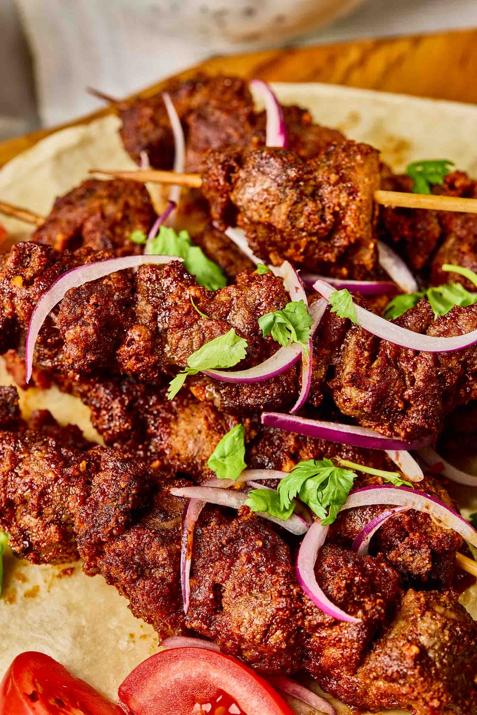 Grilled suya skewers of seasoned grilled beef topped with sliced red onions and fresh cilantro, served on a flatbread with tomato slices.