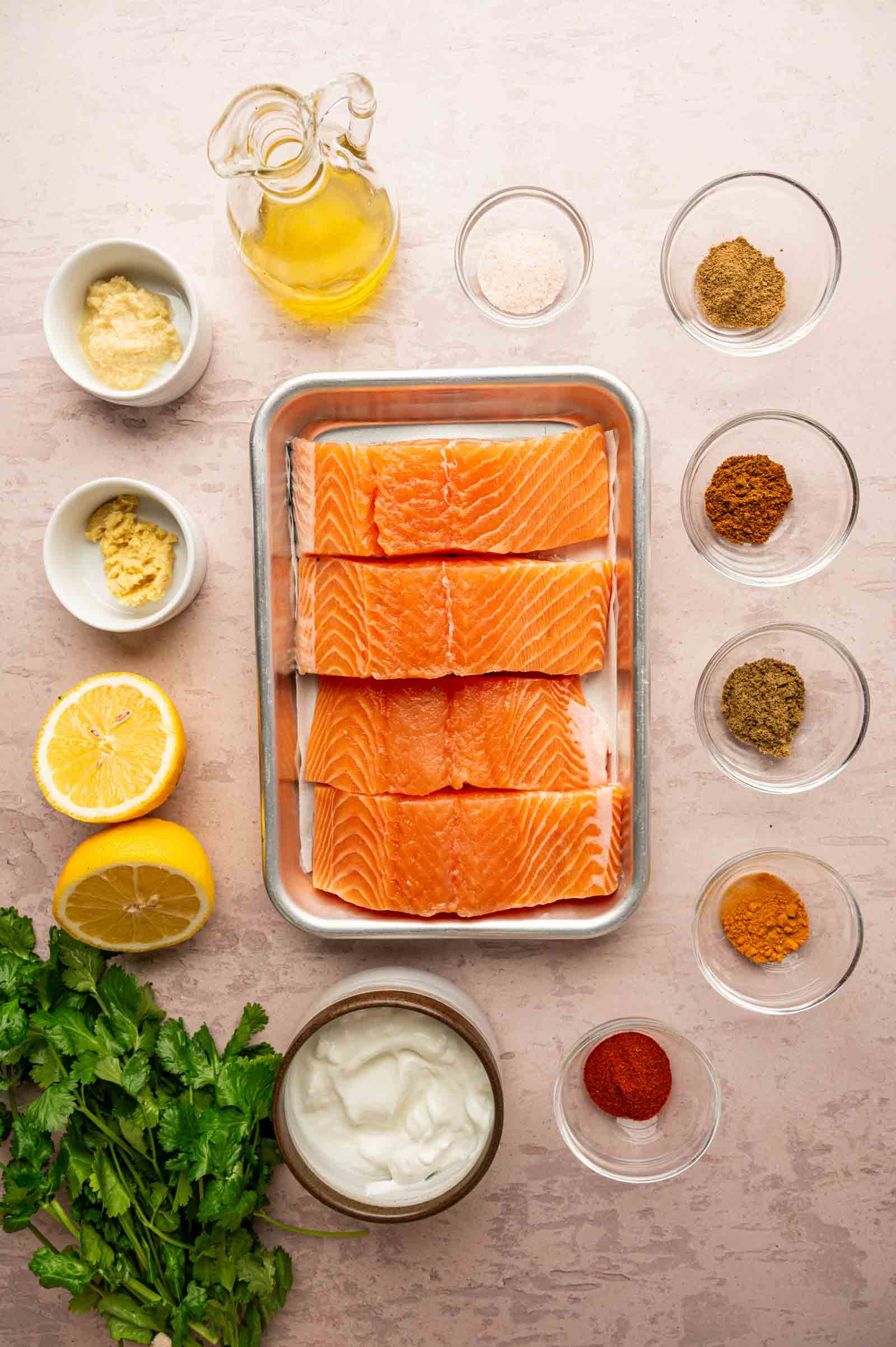 Raw salmon fillets in a tray surrounded by small bowls of spices, yogurt, mustard, oil, salt, lemon halves, and fresh cilantro on a light-colored surface.