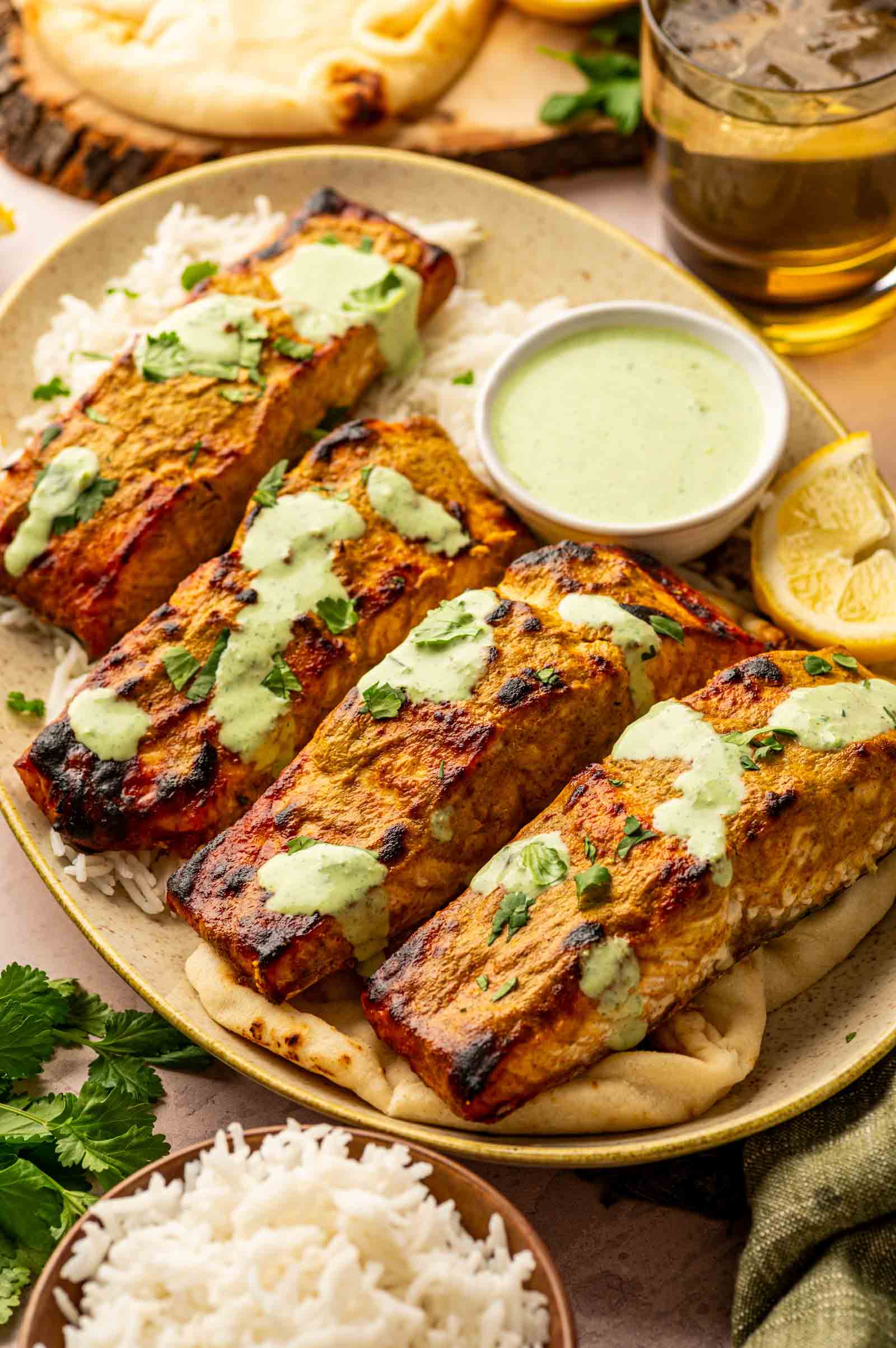 Tandoori salmon fillets with green cilantro sauce served on rice and naan, garnished with cilantro, with lemon wedges and a bowl of sauce on the side.