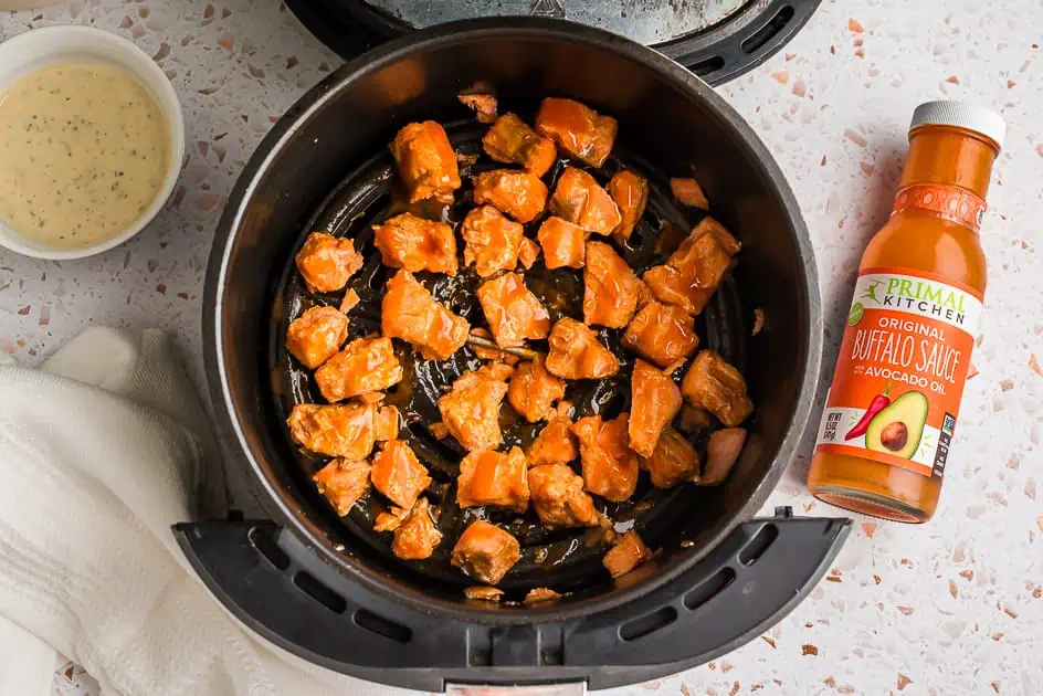 Air fryer filled with buffalo salmon bites, bottle of buffalo sauce on the side.