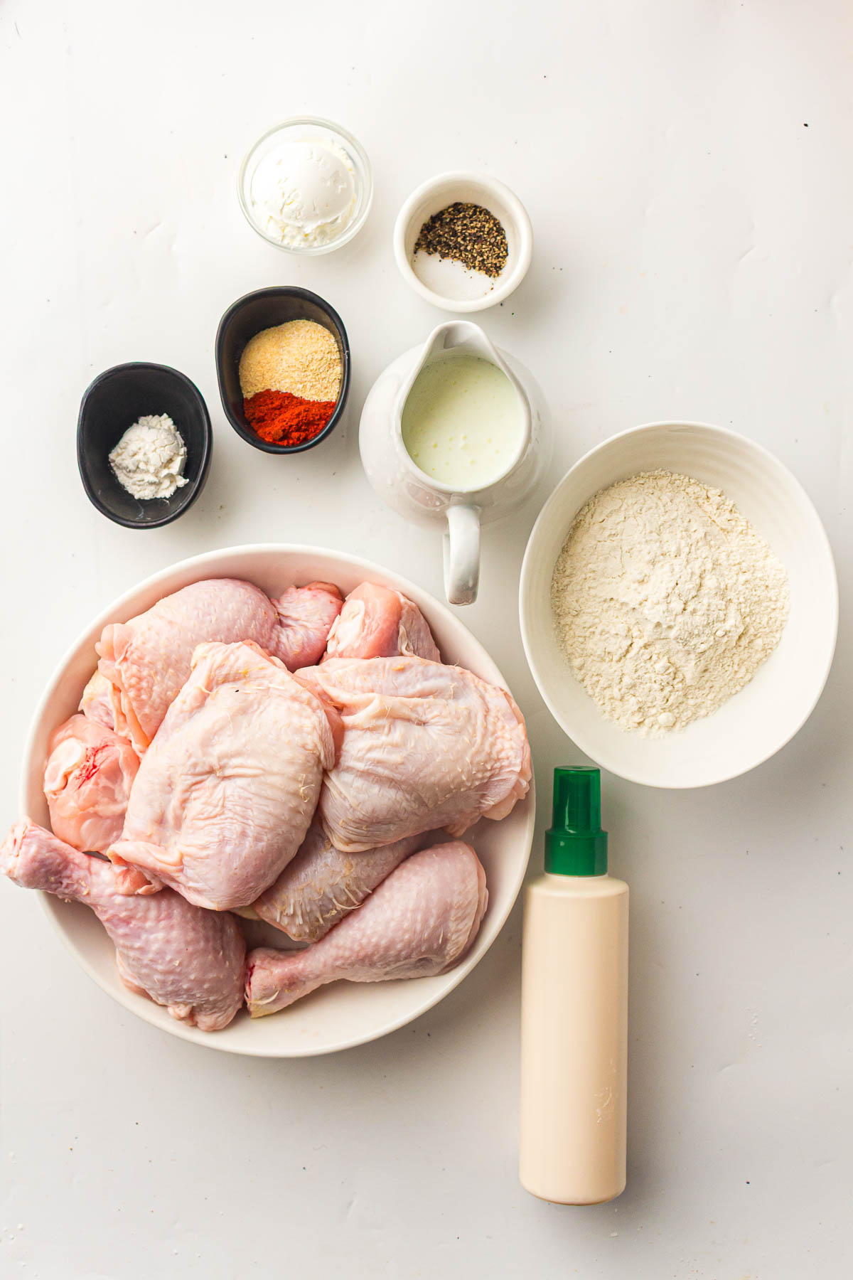 A bowl of raw chicken drumsticks surrounded by small bowls containing flour, spices, black pepper, a white liquid, and a bottle with a green nozzle on a white surface.