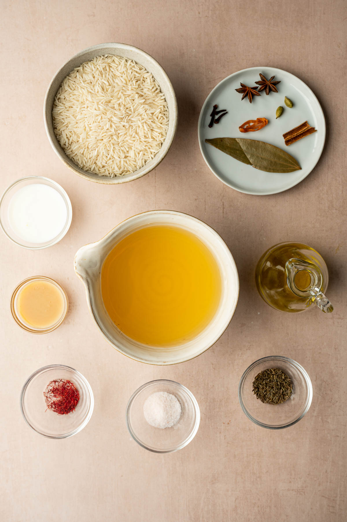 Overhead view of measured ingredients for cooking, including rice, spices, milk, oil, saffron, salt, and broth arranged neatly on a light surface.