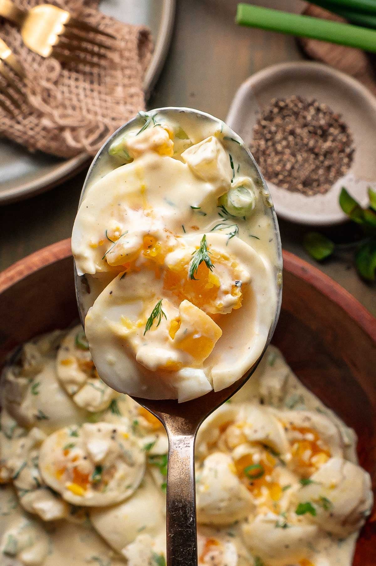 A close-up of a spoon holding a creamy broken egg salad garnished with dill, with a bowl of the same salad and a plate of black pepper in the background.