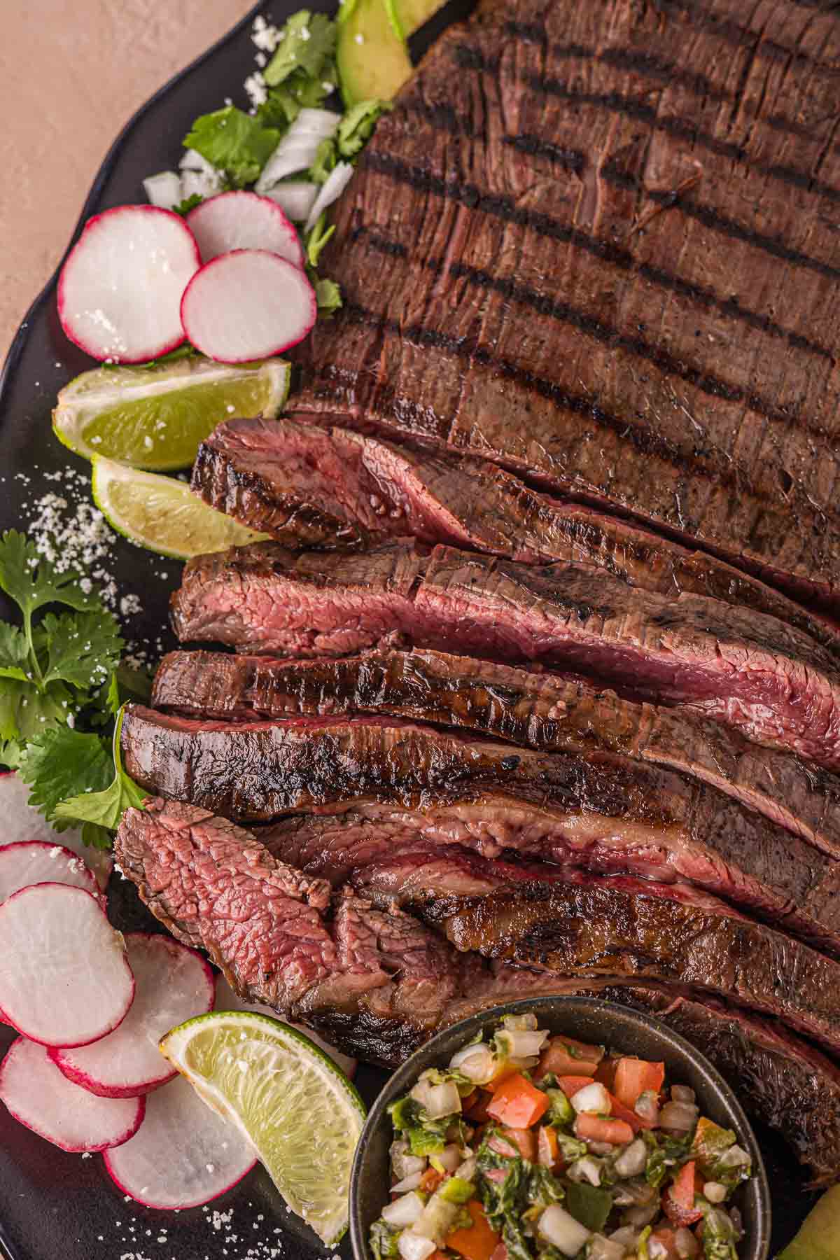 Grilled and sliced carne asada served with lime wedges, sliced radishes, cilantro, chopped onions, and a small bowl of salsa on a black plate.