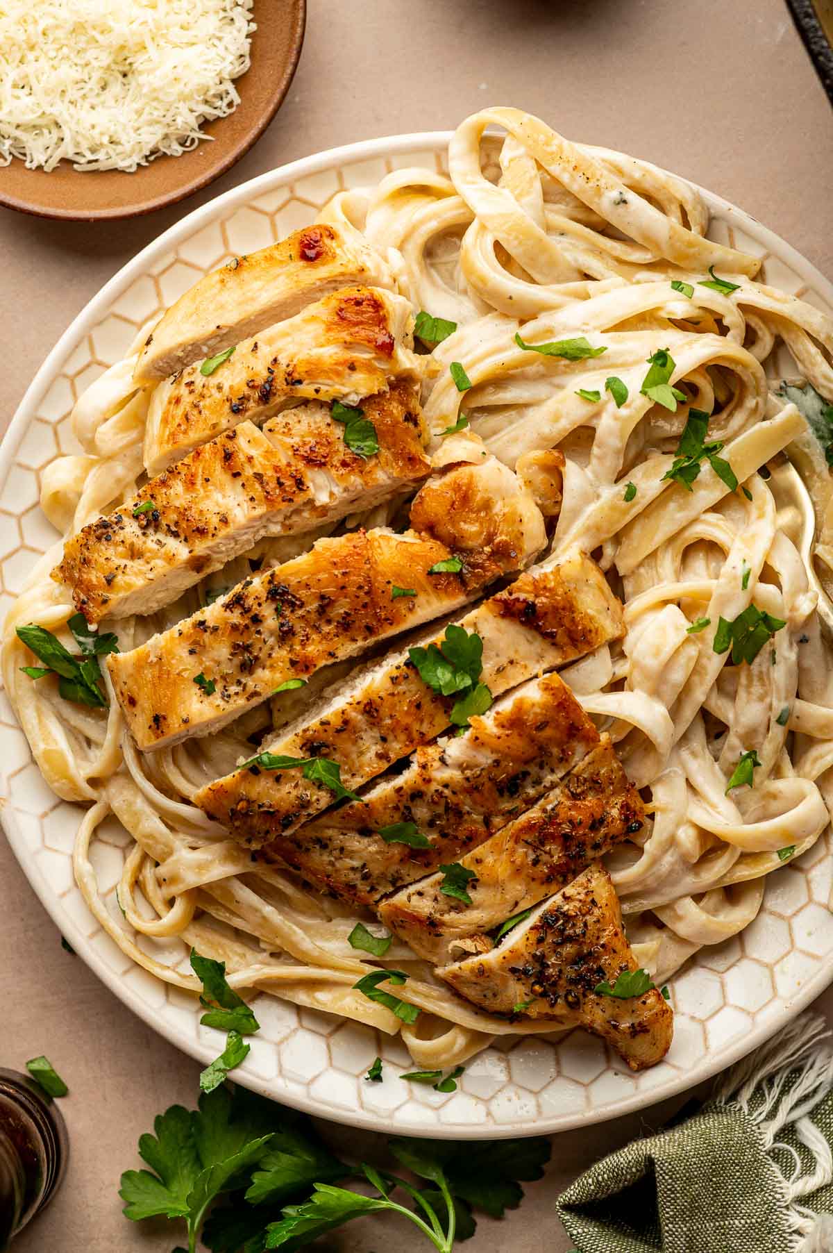 A plate of fettuccine Alfredo topped with sliced, seasoned grilled chicken breast and garnished with chopped parsley.