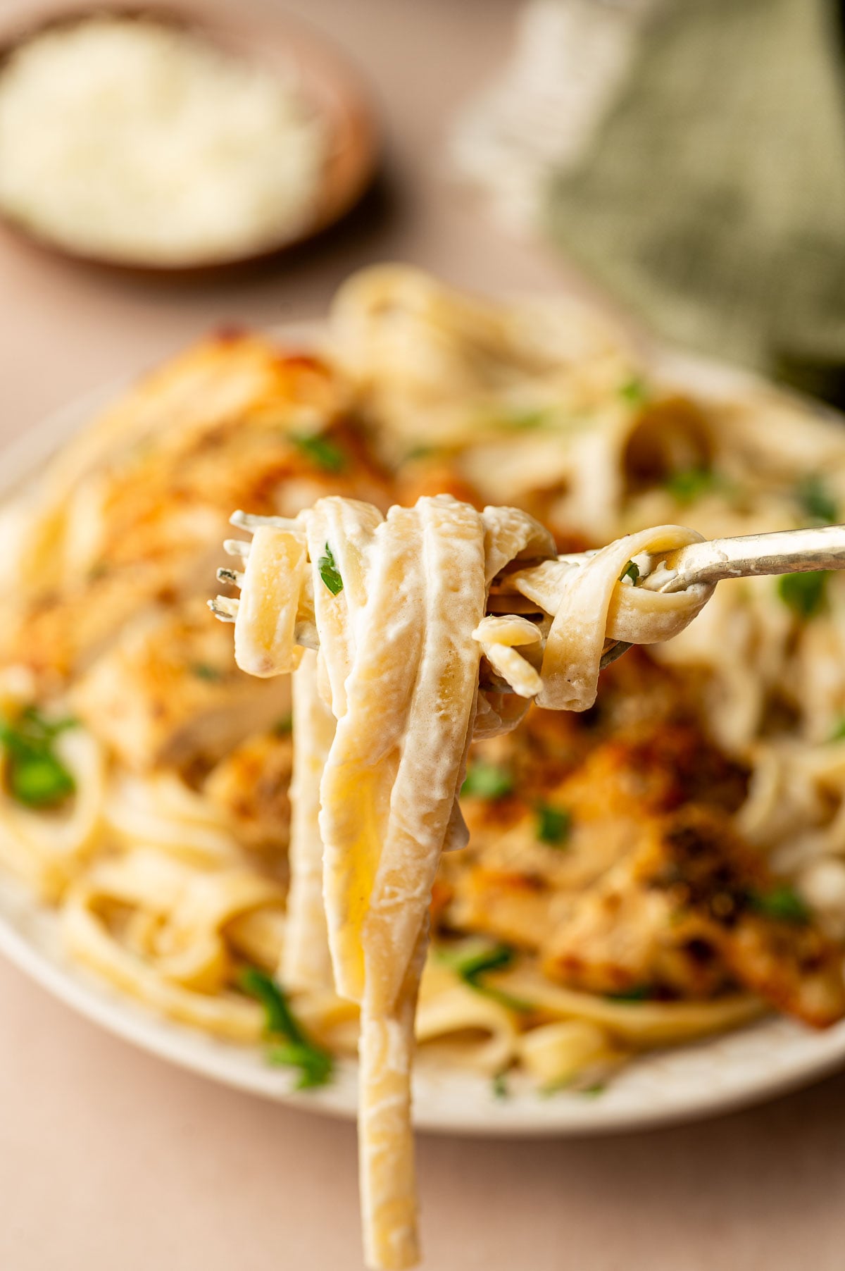 A close-up of a fork holding creamy fettuccine pasta above a plate of pasta with sauce and herbs in the background.