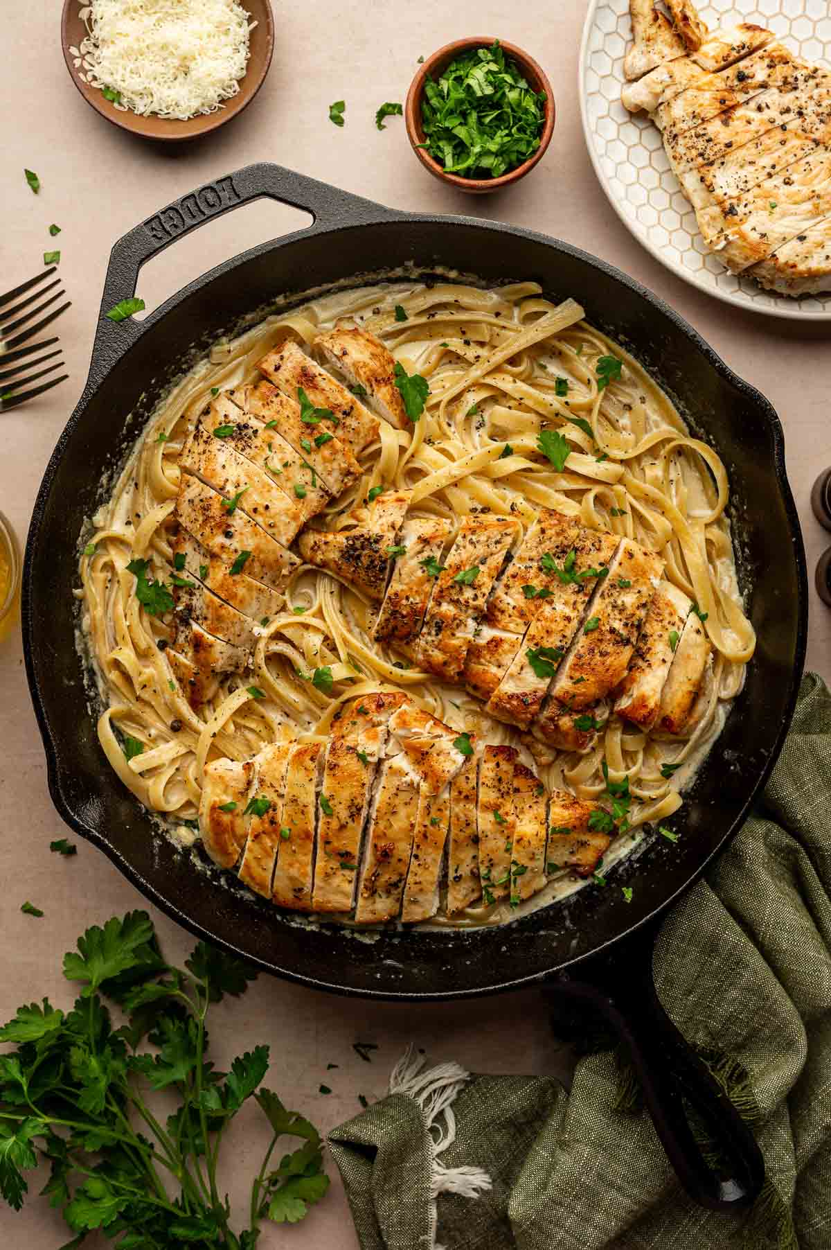 A cast iron skillet filled with fettuccine pasta in a creamy alfredo sauce, topped with sliced grilled chicken breast and garnished with chopped parsley.