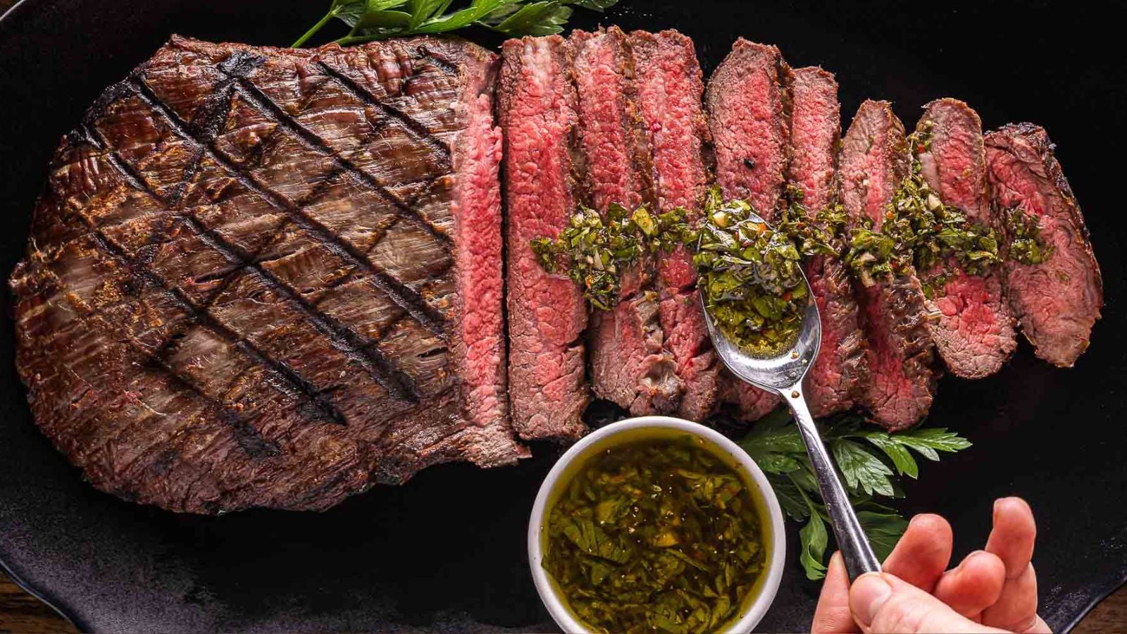 Sliced grilled steak drizzled with bright green chimichurri sauce on a wooden cutting board.