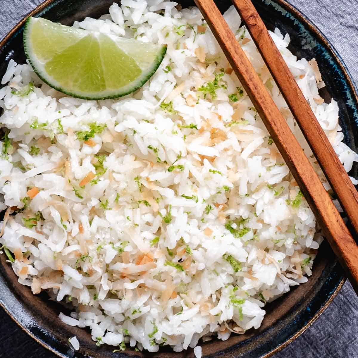 Coconut rice with lime wedges, lime zest, toasted coconut, and chopsticks in a dark bowl.