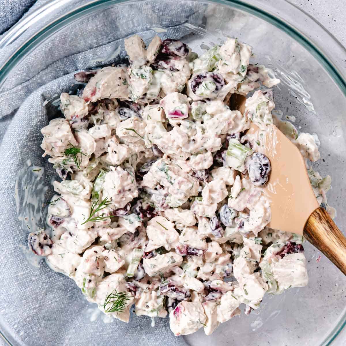 Turkey salad with dried cranberries, celery, red onion, and fresh herbs being mixed in a glass bowl.