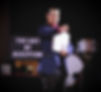 Man in a suit pointing, holding a notebook on stage. "The Art of Deception" sign behind him. Drum set visible. Focused mood.