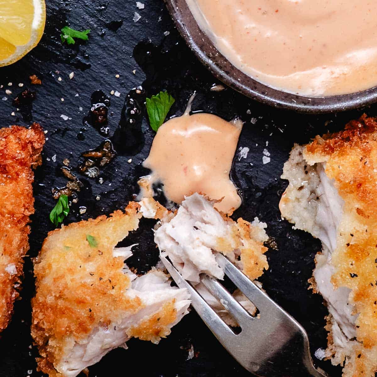 Crispy fried fish pieces with bang bang sauce, lemon, and parsley on a black slate plate