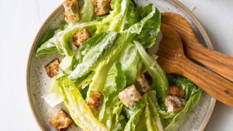 A plate of Caesar salad with romaine lettuce, croutons, and dressing is served with wooden salad tongs. A small glass jar of dressing with a striped straw sits nearby on a white surface.