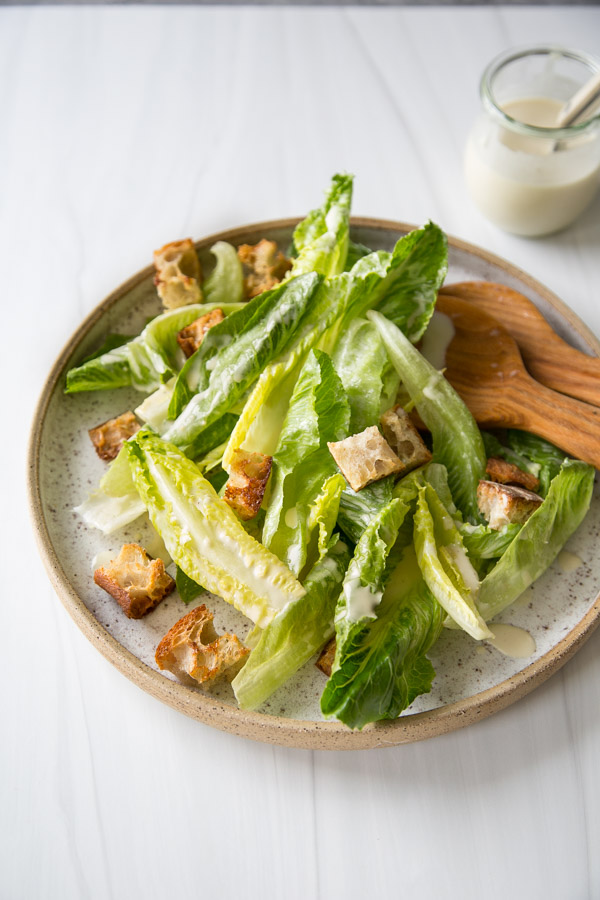 A plate of Caesar salad with romaine lettuce, croutons, and creamy dressing, served with wooden utensils on a light surface. A small jar of dressing is visible in the background.