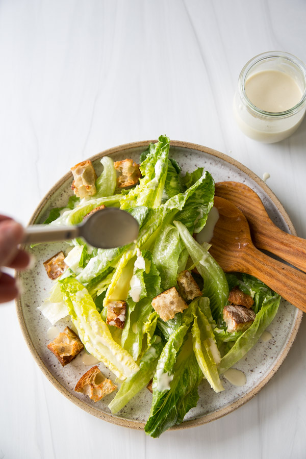 A plate of Caesar salad with romaine lettuce and croutons is being drizzled with dressing from a spoon. A wooden salad serving set and a jar of extra dressing are nearby on a white surface.