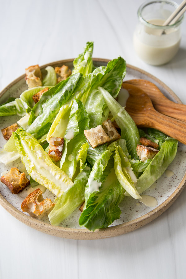 A plate of Caesar salad with romaine lettuce, croutons, and creamy dressing, served with wooden salad tongs. A small jar of extra dressing sits in the background on a white surface.