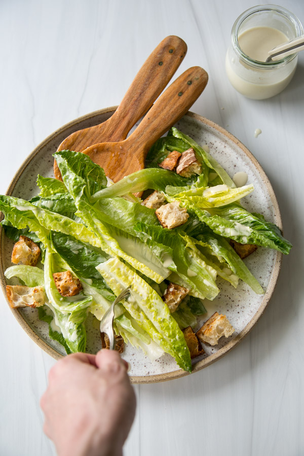 A hand holds a fork, ready to eat a Caesar salad with romaine lettuce, croutons, and dressing on a plate with wooden salad servers; a jar of extra dressing sits nearby on a white surface.