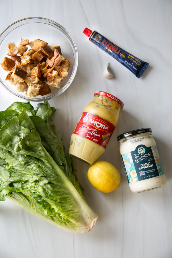 A bowl of bread cubes, a tube of anchovy paste, a garlic clove, Dijon mustard, a jar of mayonnaise, a lemon, and a head of romaine lettuce arranged on a white surface.