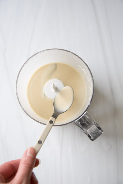 A hand holds a spoon above a food processor bowl containing a creamy, light-colored mixture on a white surface.