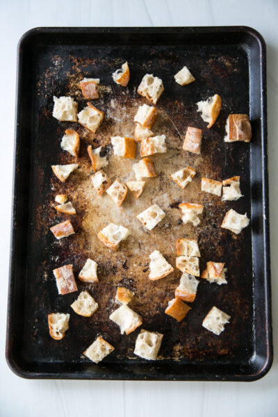 Cubed pieces of bread scattered on a dark, worn baking sheet, ready for toasting or baking, with a few crumbs visible on the tray.