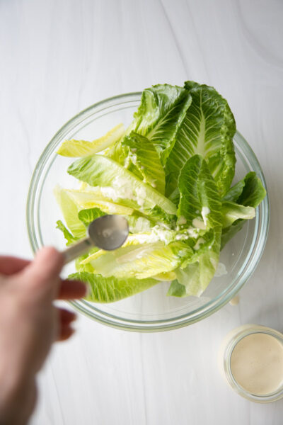 A hand holding a spoon is pouring creamy dressing over fresh romaine lettuce leaves in a clear glass bowl, with a small jar of dressing nearby on a white surface.