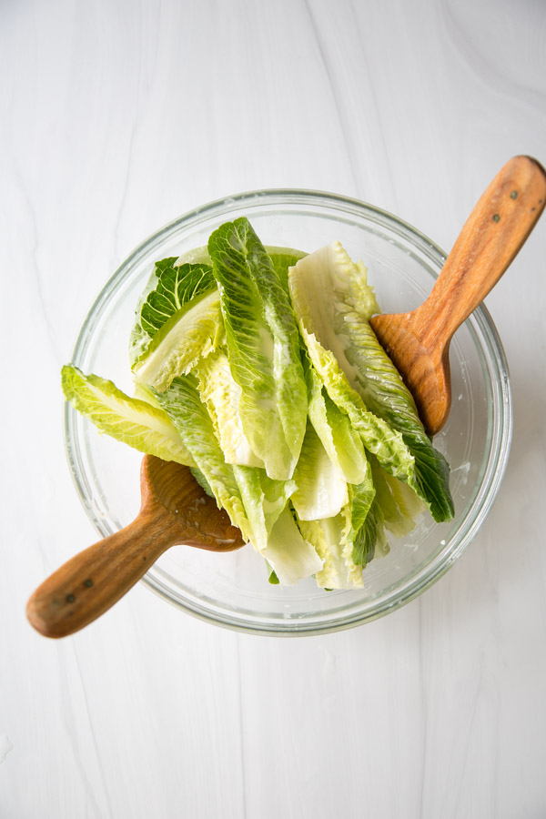 A glass bowl filled with fresh romaine lettuce leaves, with two wooden salad servers resting on the sides, viewed from above on a white surface.