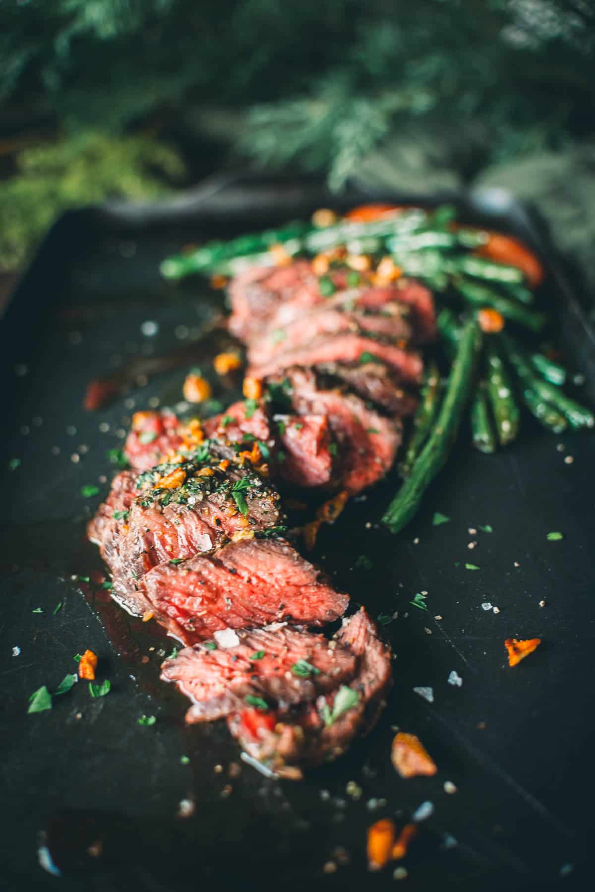 Slices of cooked beef garnished with herbs and spices on a black surface, accompanied by green beans.